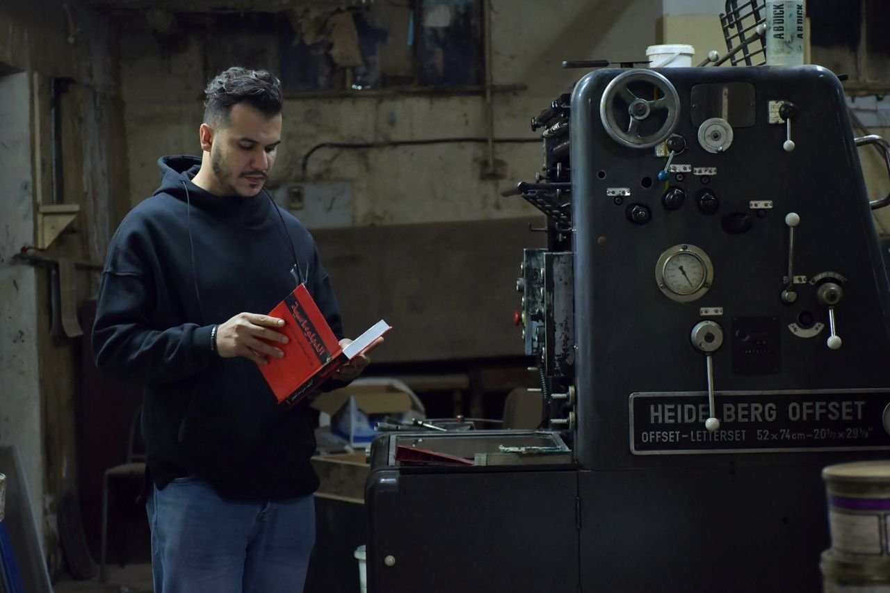 A man in a black hoodie reads by an industrial printing machine in Benghazi, Libya.