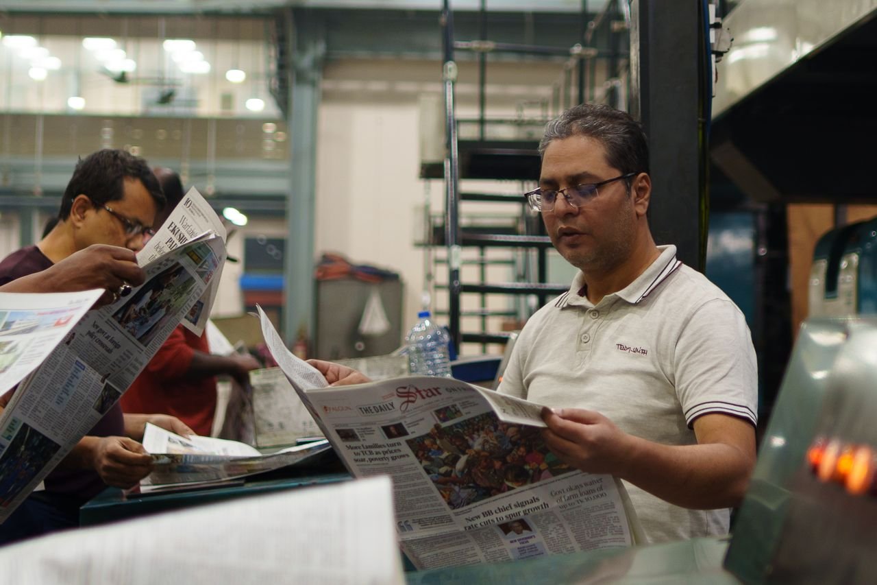 Workers at a newspaper printing press reading freshly printed copies. Focus on a man holding a newspaper.