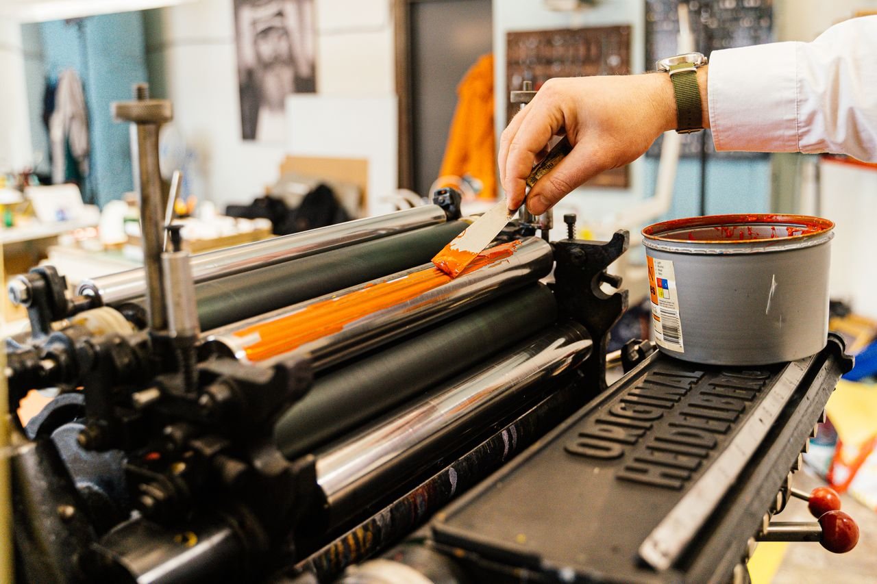 A detailed view of a hand applying orange ink to a vintage printing press roller indoors.